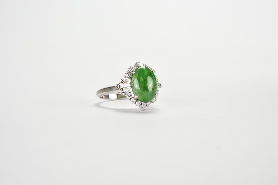 A Jadeite Cabochon and Diamond Ring: Mounted in 18K White Gold Set with an oval jadeite cabochon of rich emerald-green color and fine translucency, the stone is framed by a radiating surround of round brilliant and tapered baguette-cut