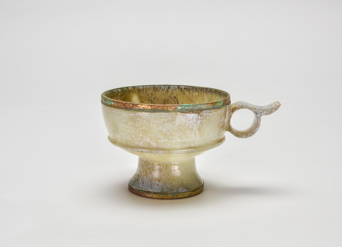 A Bronze-Mounted Jade Ear Cup with Mandarin Ducks, Han Dynasty: The vessel is carved from pale celadon jade of translucent tone, the deep rounded body raised on a spreading foot and applied to the side with an openwork ear-shaped handle terminating in an upswept p