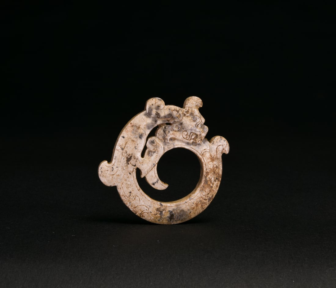 A Jade Chi-Dragon-Form Pendant, Han Dynasty: The pendant is carved in openwork as a sinuous chi dragon coiled around a large central aperture, the head rendered in relief with prominent eyes, curled horns and a hooked snout. The curving body is 