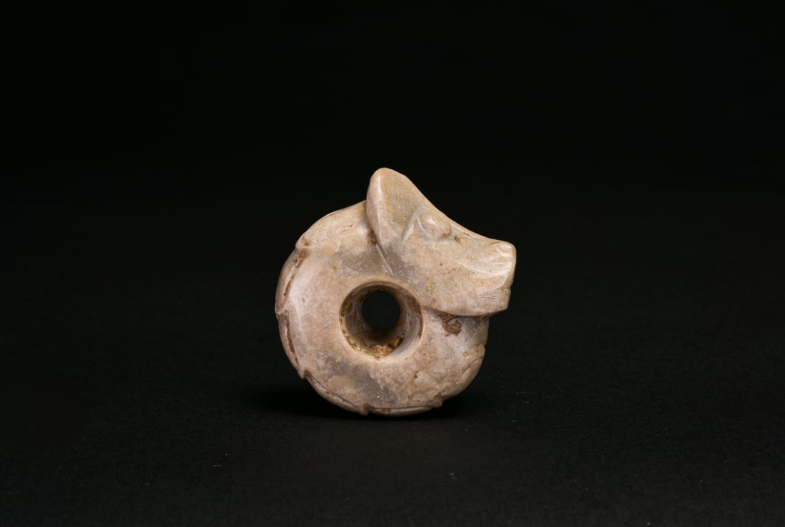 A Jade Tiger-Form Pendant. Neolithic Period: The pale beige jade is carved in the round as a recumbent tiger, its head rendered with prominent rounded eyes, upright ears, a broad muzzle and incised mouth. The compact body is articulated with fou