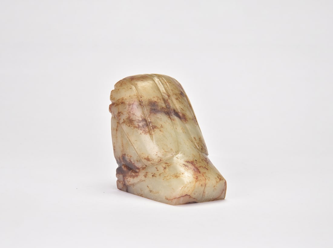 A Celadon and Russet Jade Bird-Form Ornament, Western Zhou Dynasty: The substantial ornament is carved in the form of a highly stylized bird, with a compact head and a broad, rounded body. The folded wings and plumage are delineated with long, gently curved grooves, t