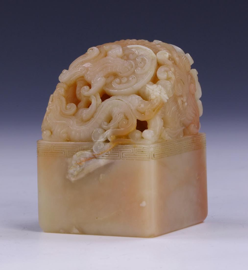 A CHINESE SOAPSTONE CARVED SEAL: A Chinese Soapstone Carved Seal: Of Rectangular Form, Rising To A Finely Carved Swirling Dragon, The Stone Of Mixed Pale Brown Tones And Mottled With Light Red Spots; Size: H: 2-1/4" (5.72cm)