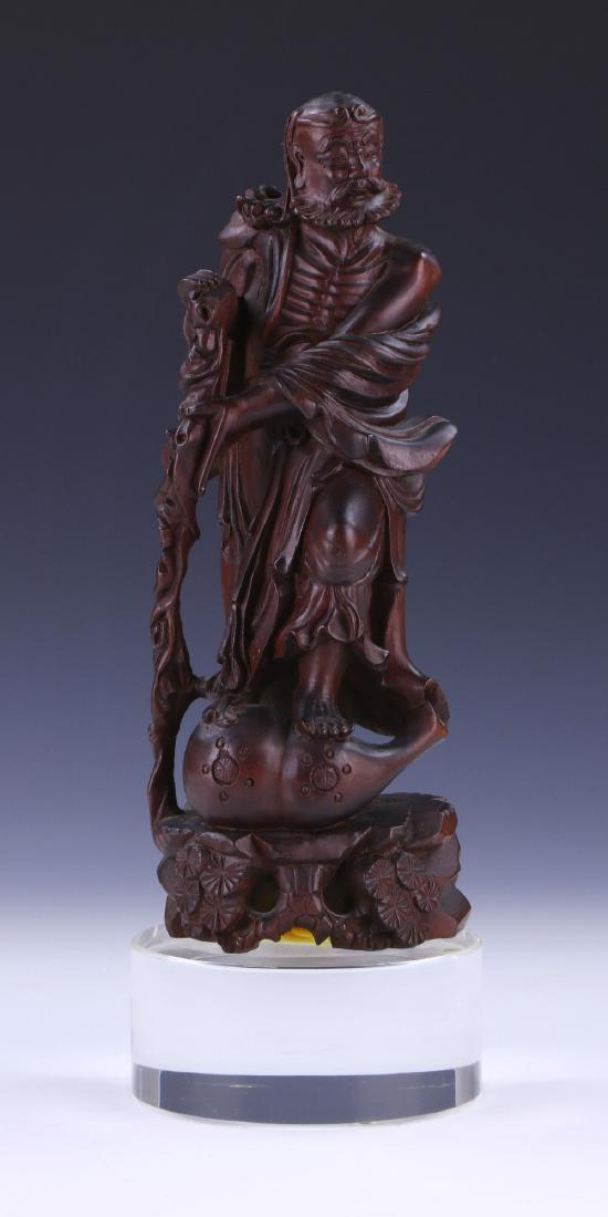 A CHINESE WOOD CARVED FIGURE: A Chinese Wood Carved Figure: Finely Carved And Shown Rising From A Cylindrical Glass Plinth, Draped in Loose Robes And Supported With A Long Staff, The Wood Of A Rich Dark Brown Tone; Size: H: 13-1/2