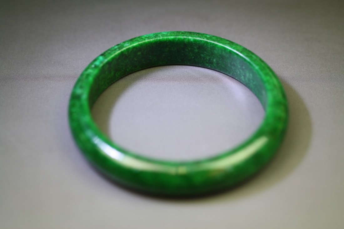 A Jadeite Bangle: A Jadeite Bangle: the well-polished emerald green jadeite flecked with areas of darker tone, measuring approximately 75mm. in diameter and 12. 8mm. in thickness; Size: D: 75mm; T: 12.8mm