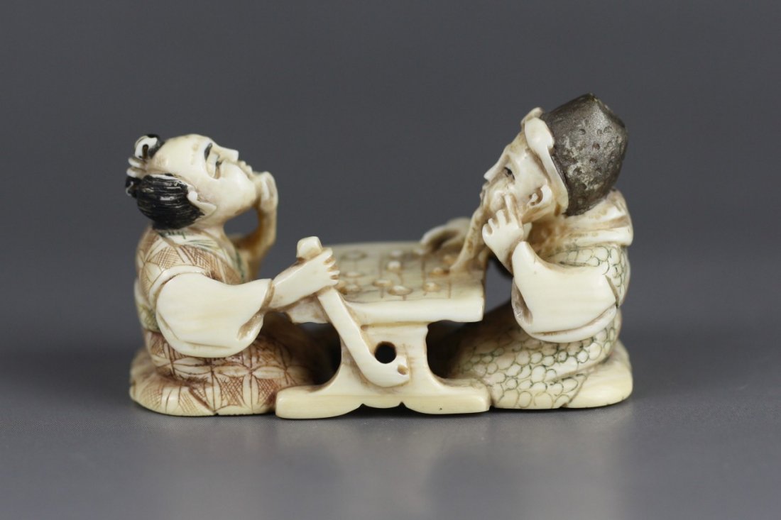 An Antique Polychrome Ivory Netsuke: Figures: An Antique Polychrome Ivory Netsuke: Two men sitting playing a board game while they have one hand on their face in a thinking expression; Size: L: 2-1/8"