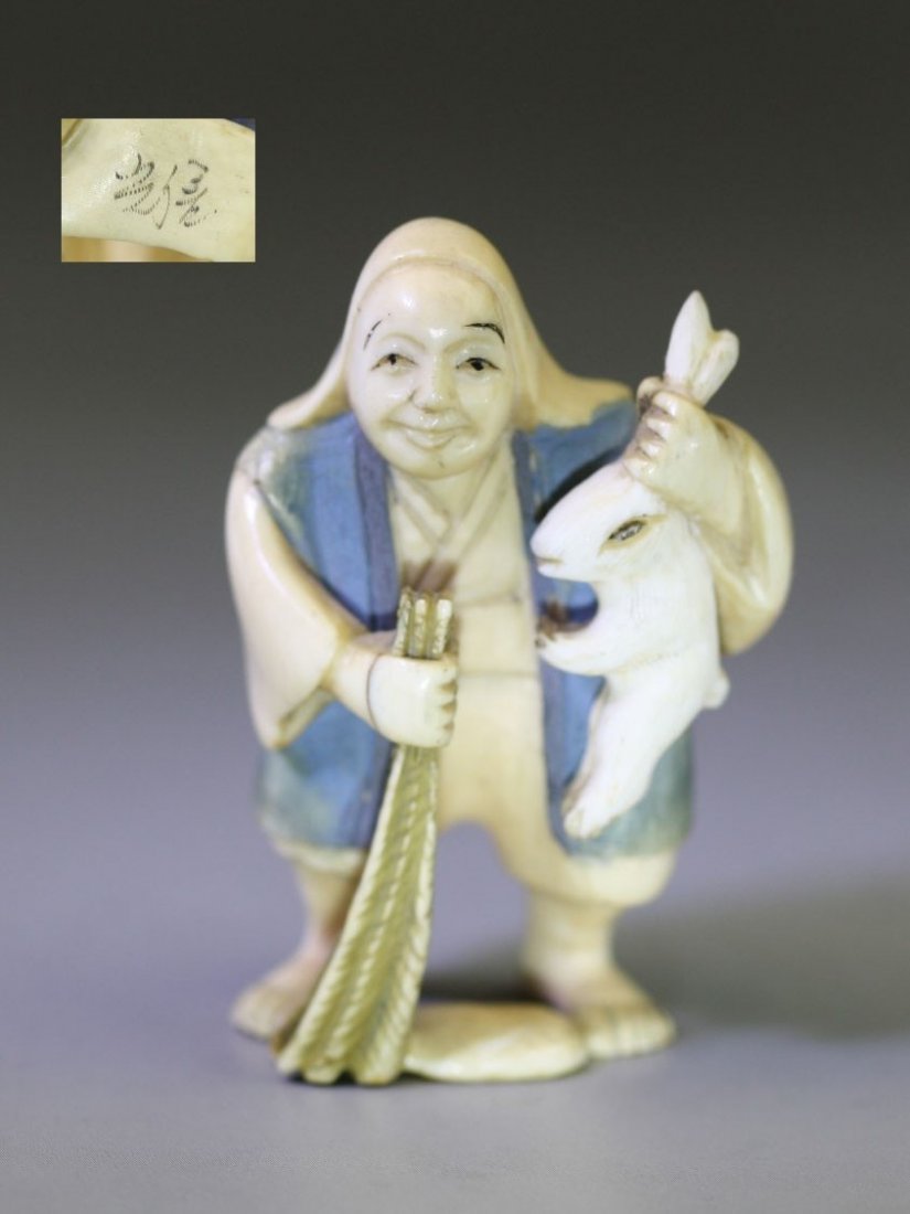 An Antique Polychrome Ivory Netsuke: Figure: An Antique Polychrome Ivory Netsuke: depicting a man standing while holding a rabbit in his left hand, and carrying a long tool in his right hand, signed on the base, 19th Century; Size: H: 2"