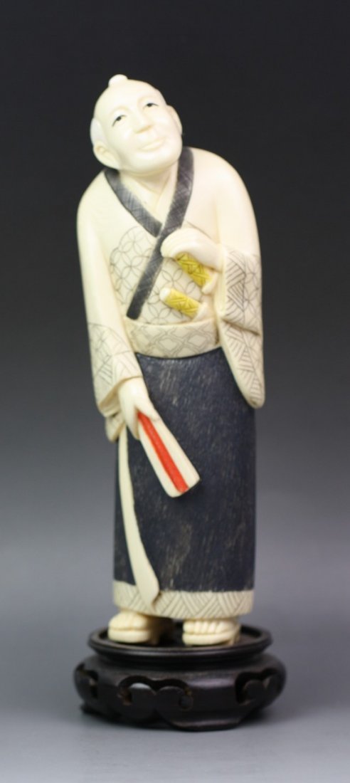 A Japanese Carved Polychrome Ivory Okimono: A Japanese Carved Polychrome Ivory Okimono: of a male figure standing in robes with two swords in his sash, holding a fan in his right hand, while right hand resting on one handle of the sword, signed