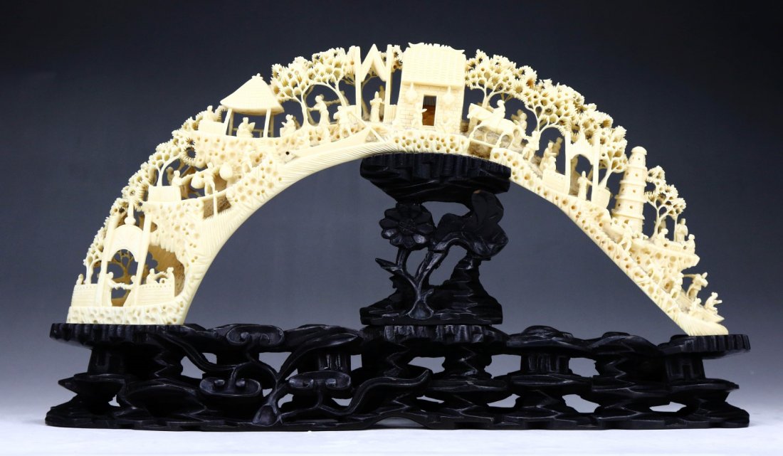 A Chinese Antique Hippo Tusk Carved Figure Group: A Chinese Antique Hippo Tusk Carved Figure Group: elaborate openwork carved in a bridge formation, numerous scenes of figures within a village and landscape, presented on a wood stand, African ivory, 