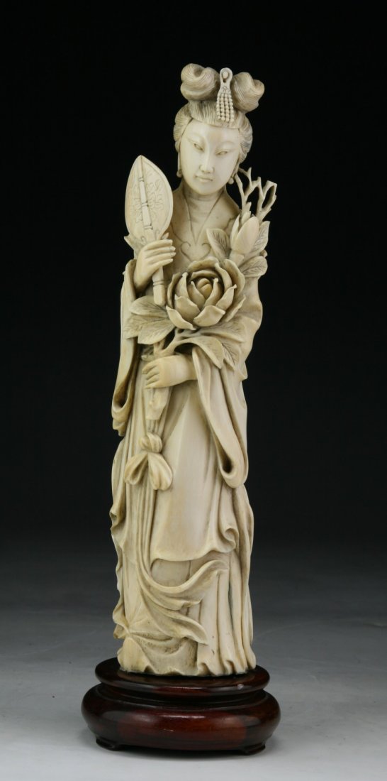 A Chinese Antique Ivory Carved Beauty: A Chinese Antique Ivory Carved Beauty: finely carved beauty standing in a long flowing robe holding a fan in one hand and blooming flowers in the other; presented on a wood stand, African ivory, Late