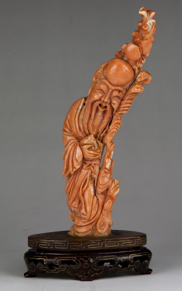A Chinese Antique Coral Carved Shoulao: A Chinese Antique Coral Carved Shoulao; finely carved as Shoulao standing in long layered robes holding a long stick with a gourd attached while holding a peach in the other hand, presented on a wood