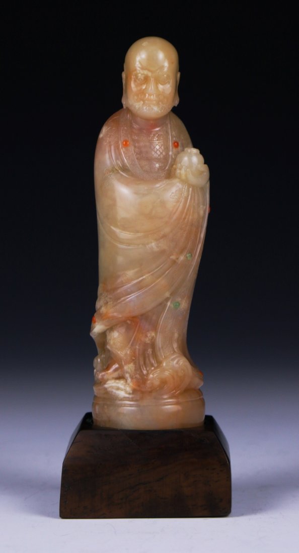 A Chinese Soapstone Carved Damo: A Chinese Soapstone Carved Damo: finely carved as Damo standing in layered robes holding a jar, inlaid with circular agate cabochons, presented on a wood stand; Size: H: 4-1/4"; (overall) H: 5"