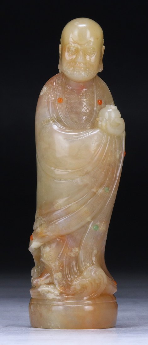 A Chinese Soapstone Carved Damo: A Chinese Soapstone Carved Damo: finely carved as Damo standing in layered robes holding a jar, inlaid with circular agate cabochons, presented on a wood stand; Size: H: 4-1/4"; (overall) H: 5"