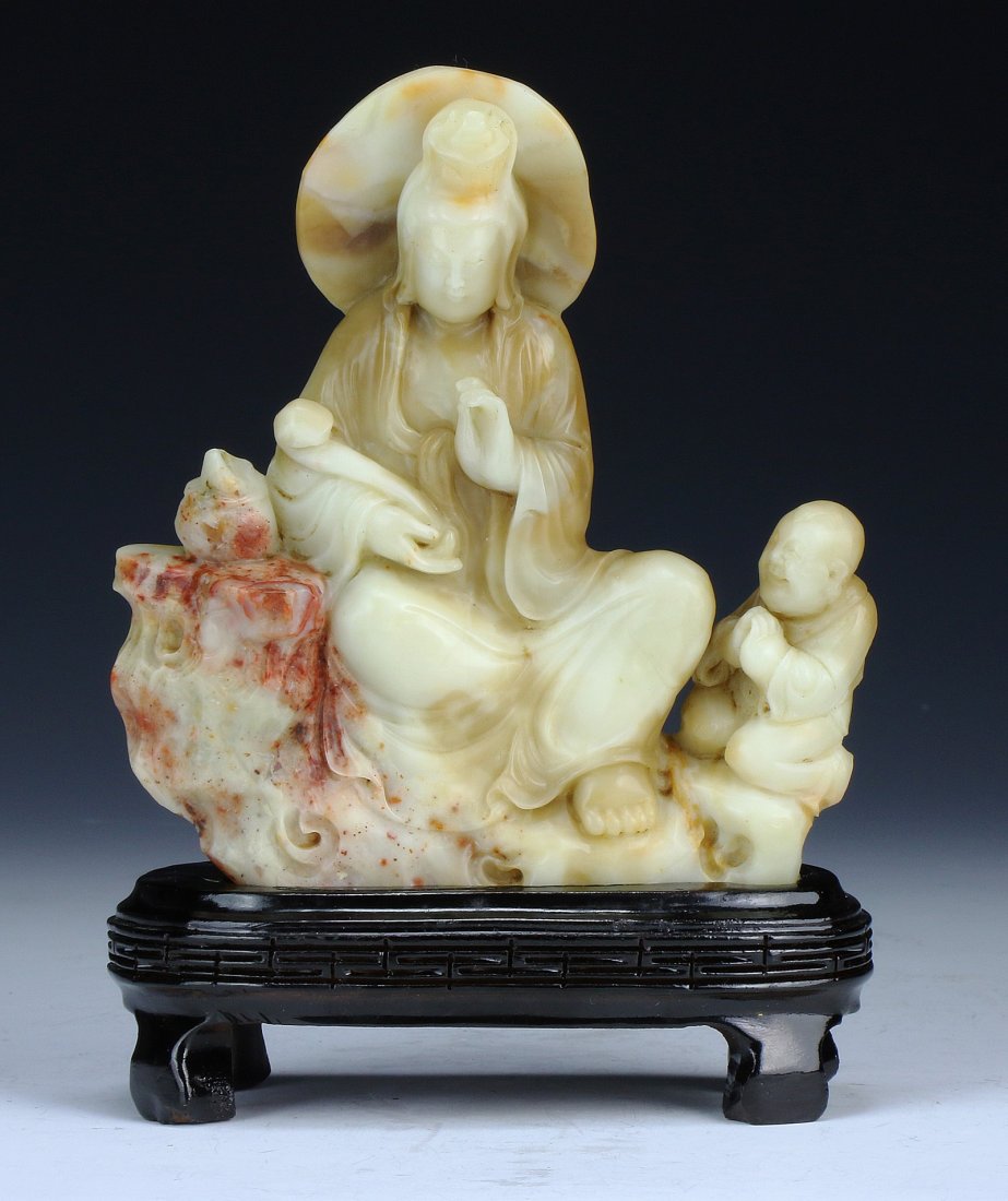 A Chinese Soapstone Carved Guanyin: A Chinese Soapstone Carved Guanyin: finely carved as Guanyin in a seated position holding a ruyi while a small child stands to her left side, the stone of creamy to honey brown tones with some red suf