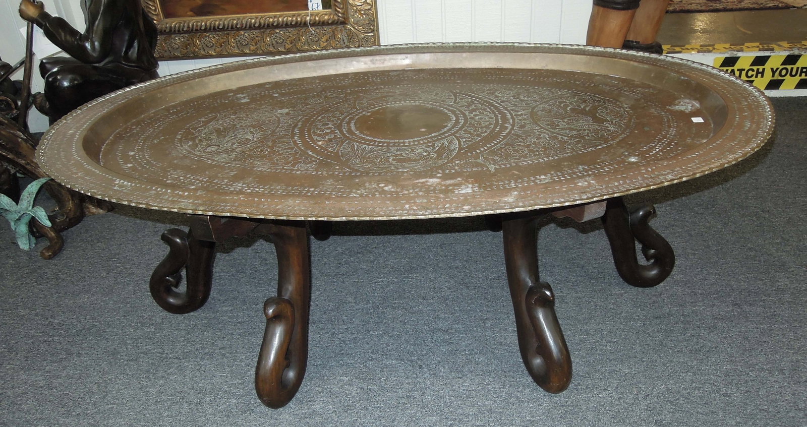 VERY RARE JOHN WIDDICOMB (GRAND RAPIDS, MI) WOODEN AND BRASS COFFEE TABLE: A heavy fitted wooden base supports an oval vintage ornate brass top. Original Widdicomb plaque on base. 17" high, 4' wide, 32" dia.