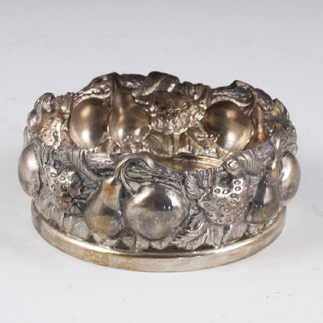 Hzorfim Sterling Wine Bottle Coaster: DESCRIPTION: A Hzorfim sterling silver wine bottle coaster. Features a rotund form embellished with repousse fruits and winding foliage. Stickered with a Hzorfim tag along the underside. CIRCA: 20th c