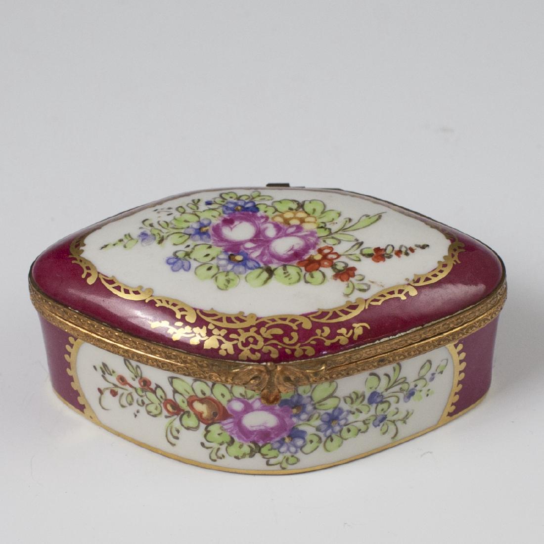 Limoges Sevres Style Porcelain Trinket Box: DESCRIPTION: A Limoges, Sevres style, porcelain trinket box. Features hand painted floral motifs enveloped by gilded scrolled rococo floral motifs and a maroon red base hue.Finished with a dore bronze