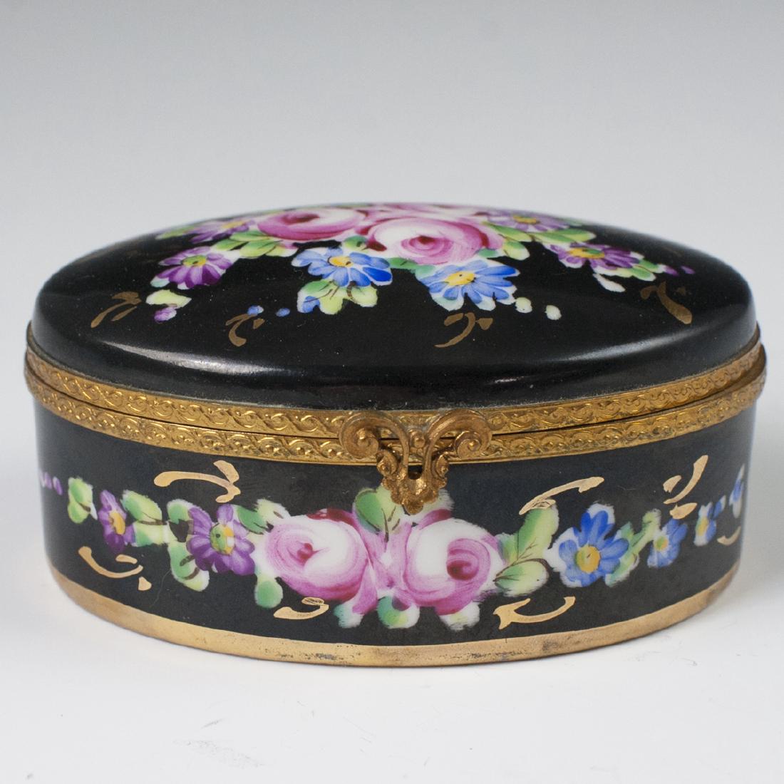 Limoges Porcelain Dresser Box: DESCRIPTION: A Limoges porcelain dresser box. Features an overall black hued base hue, embellished with floral motifs and gilded trims. Finished with a gilt metal lining and a scrolled rococo clasp. M