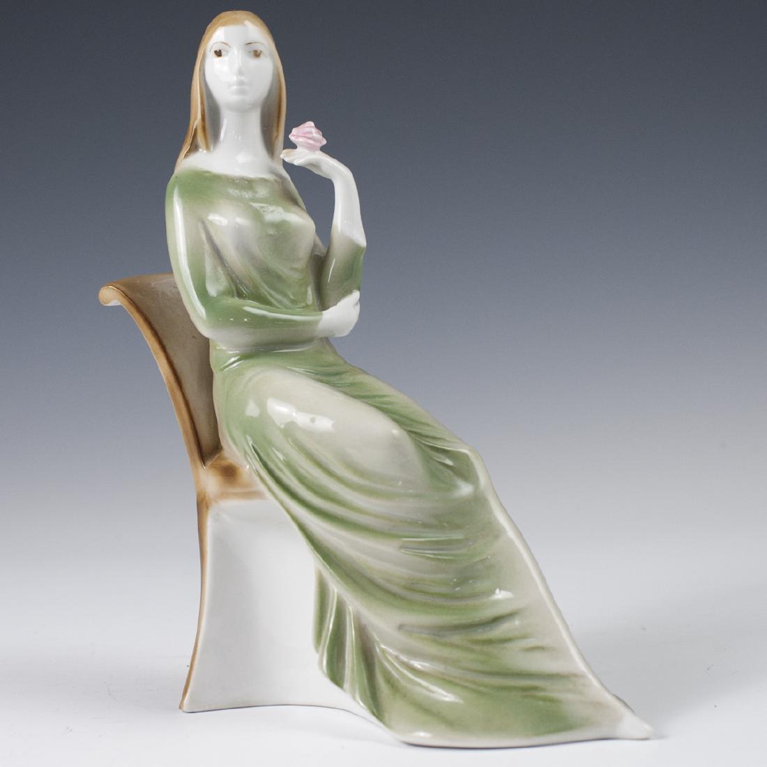 Zsolnay Porcelain Figurine: DESCRIPTION: A Zsolnay porcelain figurine of a woman wearing a dress, seated in a chair, holding a rose in her left hand. Finished in a gloss glaze and marked along the underside with a Zsolnay mark.
