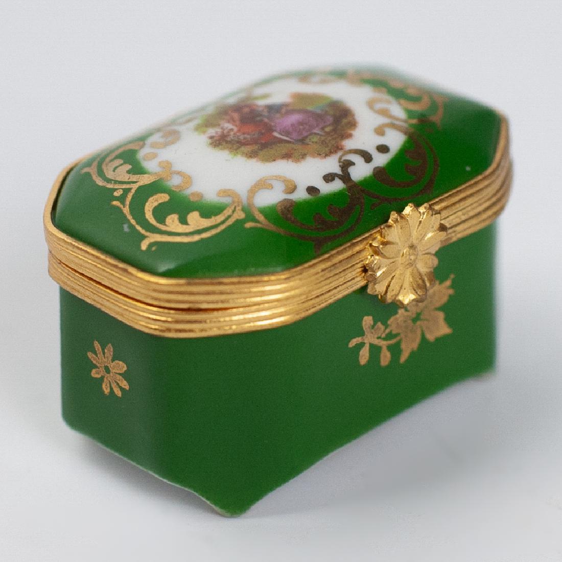 La Reine Limoges Porcelain Pill Box: DESCRIPTION: A La Reine, Limoges porcelain pill box. Features a green base tone, embellished with a center scene on the lid of a man courting a woman scene. Finished with gilded floral motifs and a fl