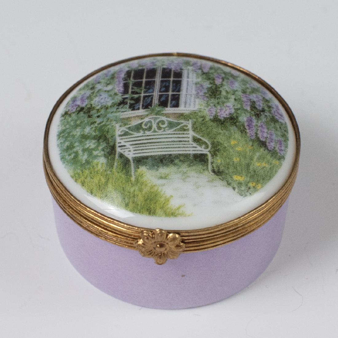 Limoges Porcelain Pill Box: DESCRIPTION: a Limoges porcelain pill box. Features a rotund form, embellished with a lavender base hue and a lid garnished with a scene of a bench in a garden backed by a window. Finished with a gilt