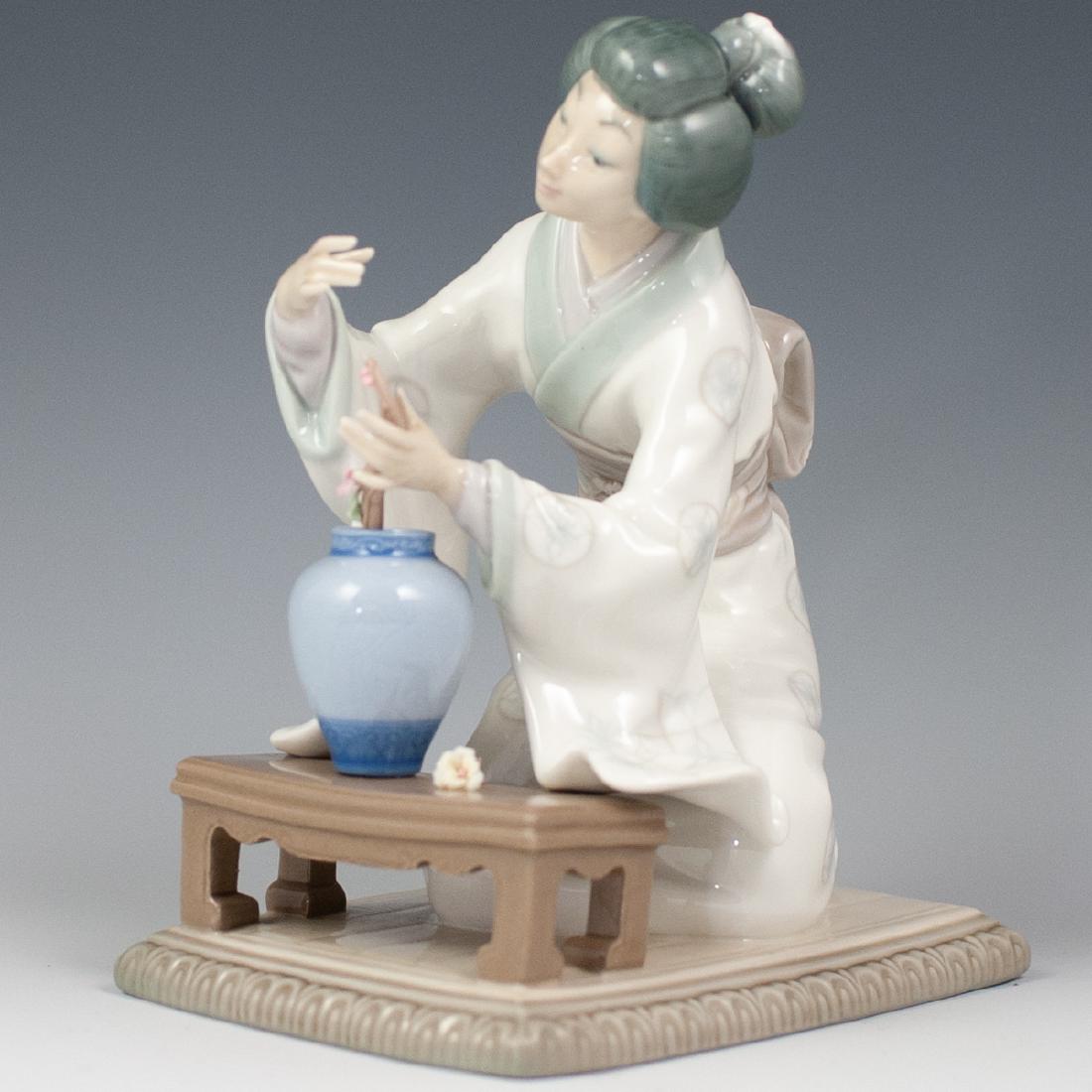 Lladro "Japanese Girl Decorating": DESCRIPTION: Lladro porcelain figurine titled" Japanese Girl Decorating". Features a Japanese girl in a white kimono, kneeling next to a table and decorating a branch with flowers. Finished with a glo