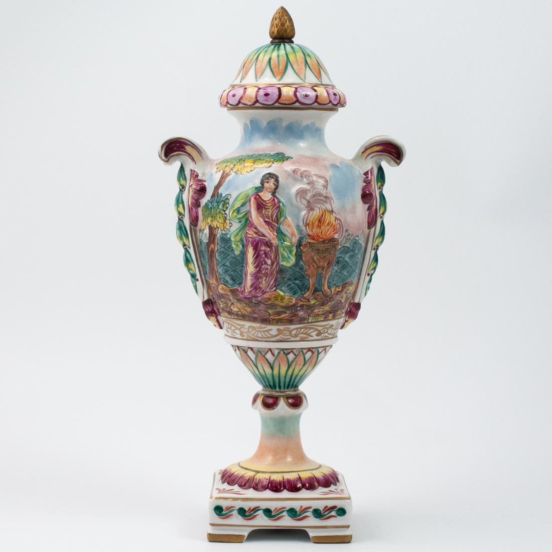 Capodimonte Porcelain Urn: DESCRIPTION: A Capodimonte porcelain urn decorated on both sides with scenes of Greco-Roman figures in multicolored hues. Flanking each side are scrolled handles embellished with hanging floweres, com
