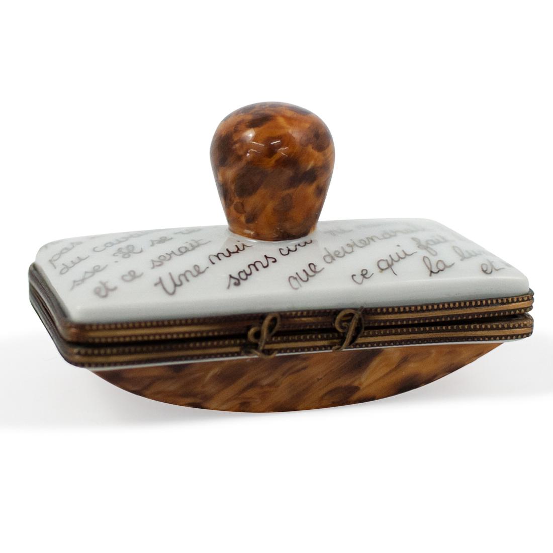 Limoges Porcelain Blotter Trinket Box: DESCRIPTION: A Limoges porcelain pill box in an overall blotter motif features written motifs along the lid with light brown hued accents. Completed with brass lining and an "LD" Clasp, marked along t
