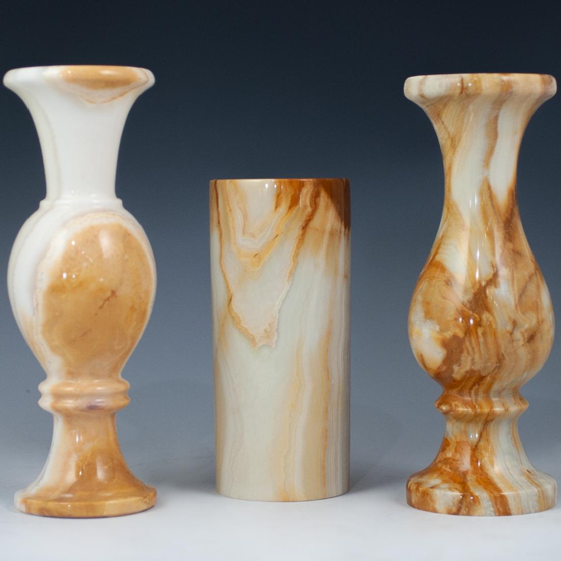 Three Alabaster Vases: DESCRIPTION: Three alabaster vases, one cylindrical vase and two trumpet vases. All three feature similar beige and white colors with splashes of honey hues. Sightly different in shape and size. CIRCA