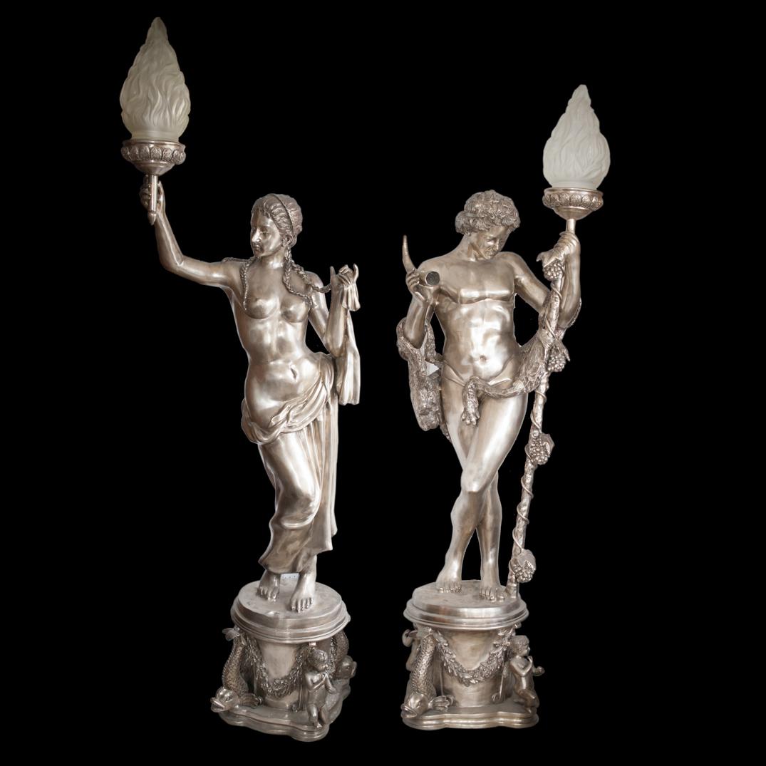 Palace Size Silver Bronze Figural Torchieres: DESCRIPTION: Pair of palace size silver bronze statue torcheres of a Greco-Roman man and woman. The man is seen holding a torch garnished with grape vines in his left hand and a horn in his right whil