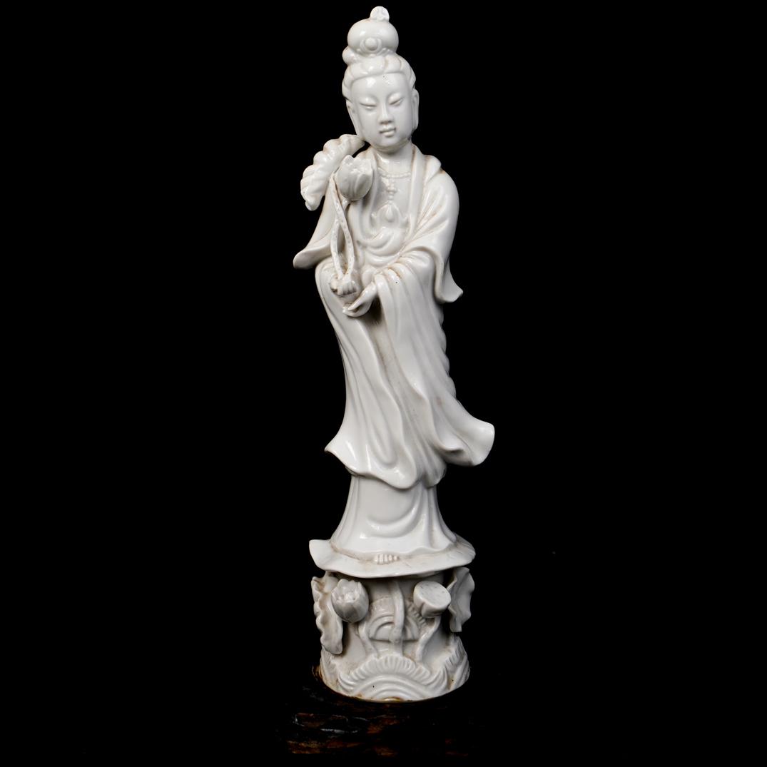 Chinese Blanc De Chine Guanyin: A antique Chinese blanc de Chine figure of a standing Guanyin, atop a verdant base with winding vines and figs. She is seen dressed in regal garments, wearing a necklace, and wielding two flowers in h