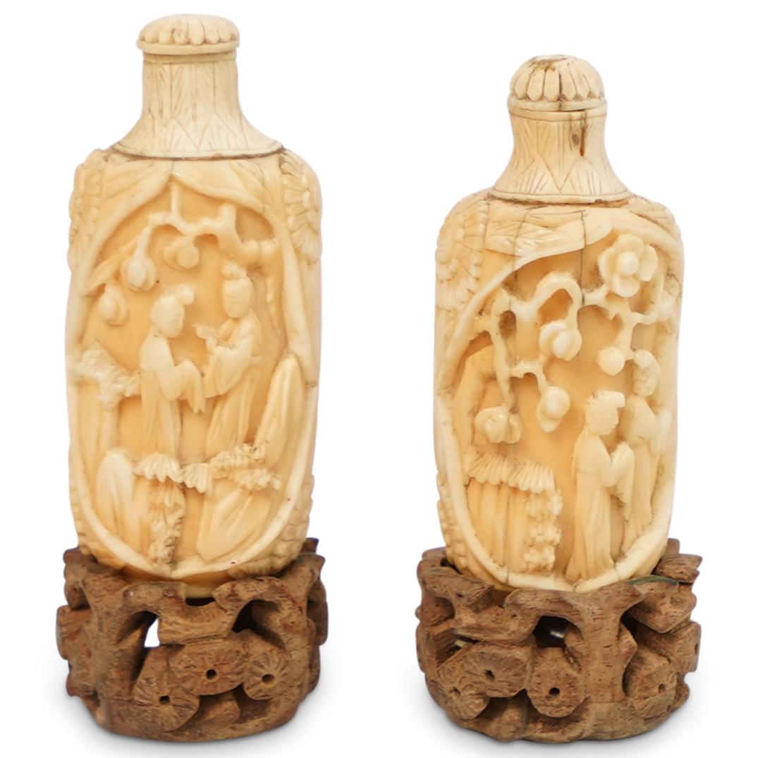 (2 Pc) Bone Carved Snuff Bottles: DESCRIPTION: (2 Pc) Antique Chinese hand carved bone snuff bottles. Features carved figural scenes under a plum blossom tree motif, finished with bamboo motif necks and flower petal tops. Rests on woo