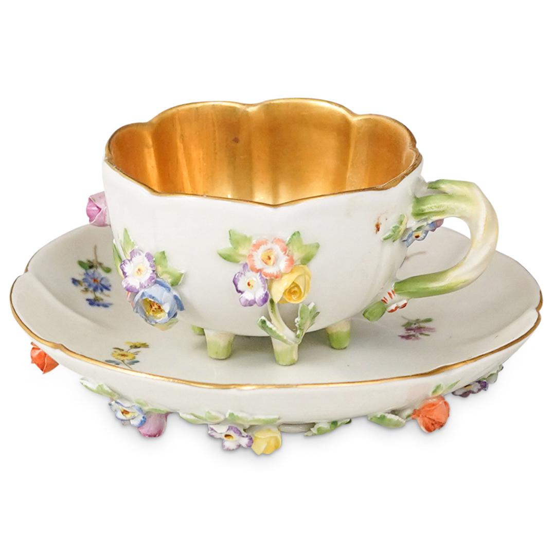 Antique Meissen Porcelain Teacup and Saucer: DESCRIPTION: An antique porcelain teacup and saucer decorated with raised floral decorations and multicolored floral motifs, accented with vine handles and gilt trims. Marked along the underside with 