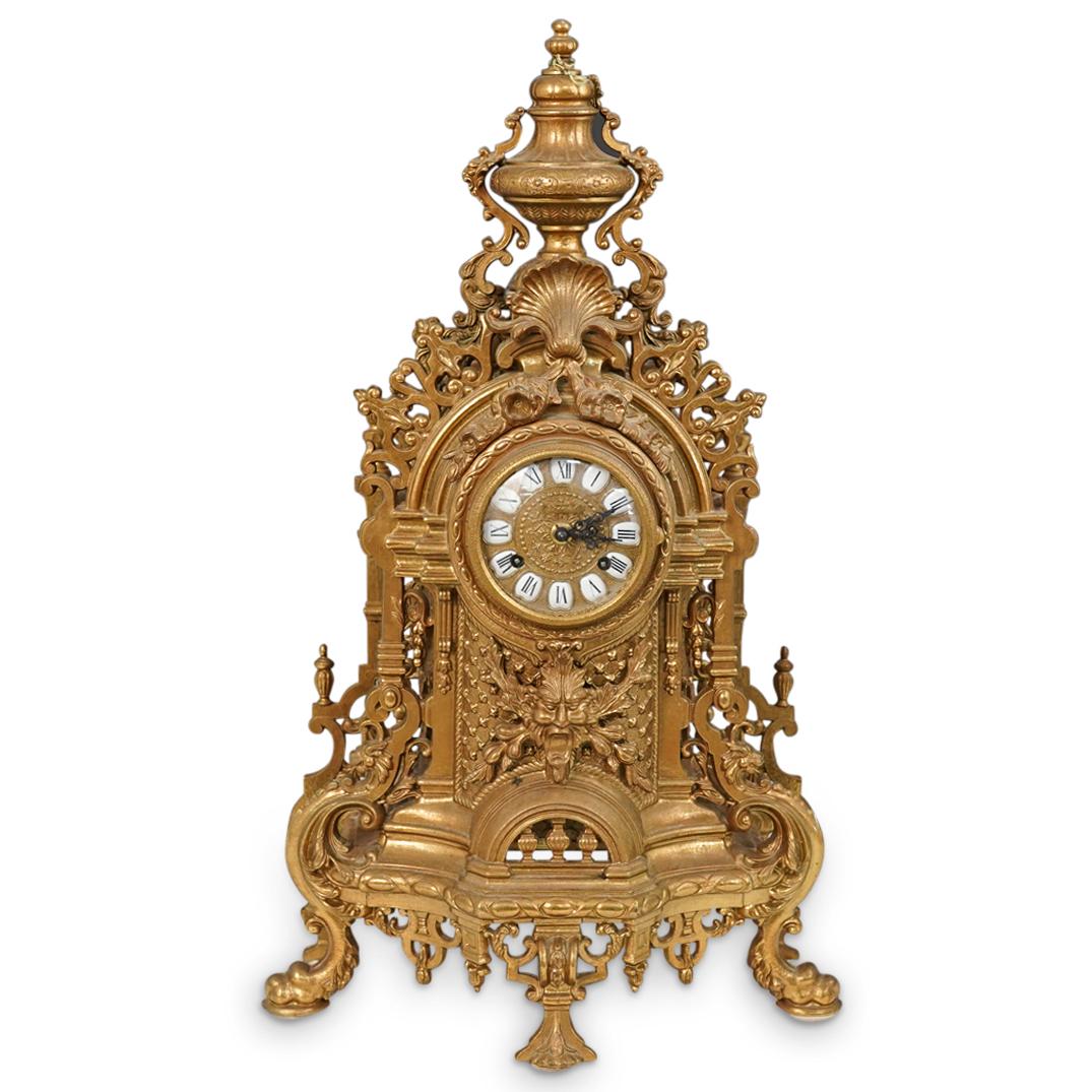 Ornate Brass Imperial Clock: DESCRIPTION: An ornate brass Imperial clock featuring an openwork case with figural masks, scrolling crests, and floral wreath details throughout. Designed with a black Roman Numeral painted enamel fa