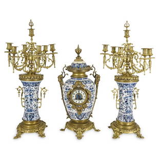 (3 pc) Delft-style Ceramic And Brass Clock With: DESCRIPTION: Delft-style porcelain and brass urn-shaped clock with matching candelabras. Porcelain parts feature a floral and avian motif similar to Royal Delft's Astonia design. Brass on clock