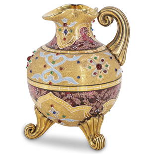 Moser Jeweled Glass Pitcher: DESCRIPTION: A Moser glass pitcher features in a Cranberry hued ground, decorated with raised gold enameling and accented with multicolored enamel and jeweled accents. Sits atop three raised