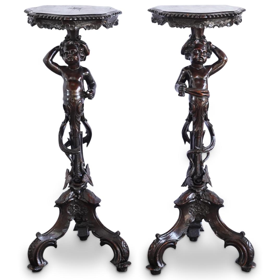 Pair Of Carved Wood Tripod Putto Columns: DESCRIPTION: Two mirrored, tripod, carved wood platforms, each featuring a stem composed of a putto bearing the top on his head and surrounded by open-work foliage designs. Piece is embellished betwee