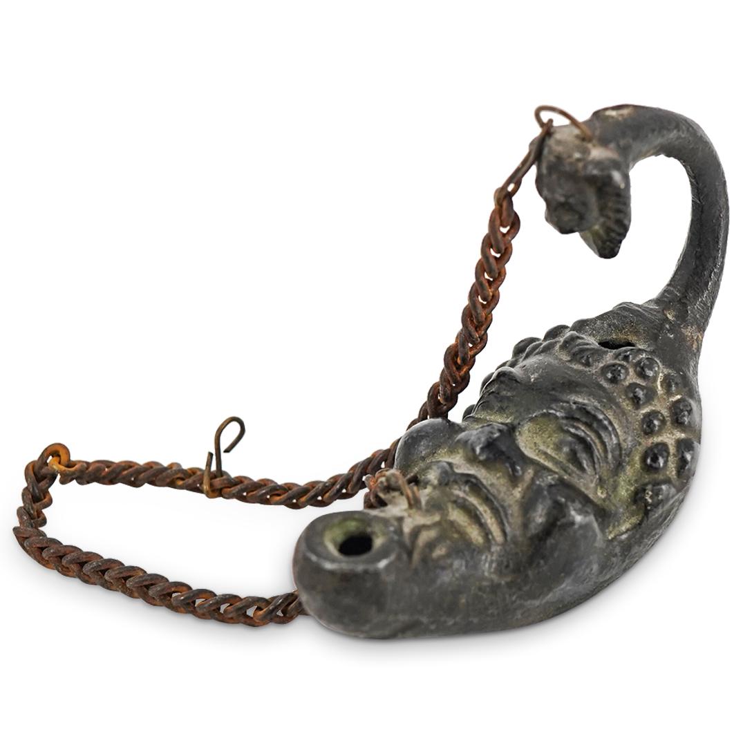 Ancient Roman Bronze Face Hanging Oil Lamp: DESCRIPTION: Ancient Roman bronze hanging oil lamp presenting a black tone throughout and a crescent form face with details of hair, eyes, a nose and lips. CIRCA: Possibly ancient. We can not guarante