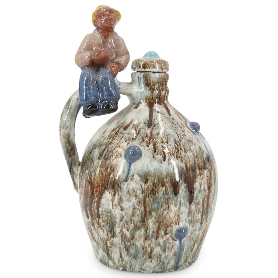 Glazed Pottery Handled Wine Bottle: DESCRIPTION: Glazed pottery handled wine bottle depicting a seated boy figure atop the handle arch. With blue, grey and brown dripping glaze finish and marked with AS hallmark. With matching lid. CIRC