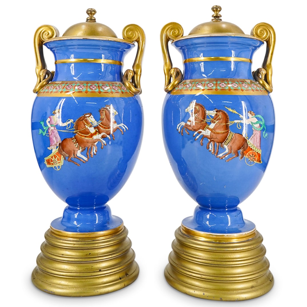 Pair Of European Neo Classical Lidded Porcelain Urns: DESCRIPTION: Two matching antique lidded porcelain urns, each featuring a neo-classical imagery depicting a goddess in a chariot opposite a medallion emblazoned with her visage. Each piece sports gilt