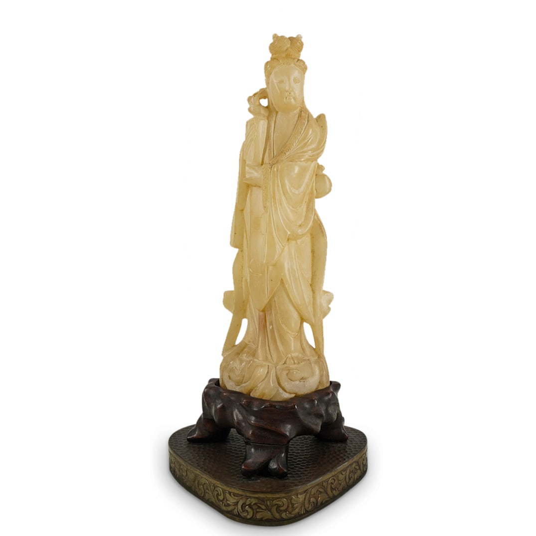 Chinese Carved Soapstone Guan Yin: DESCRIPTION: Carved soapstone sculpture depicting Guan Yin bearing an effect in her hands at her right while wearing robes and standing atop rolling waves. Figure is mounted on a carved wood base surm