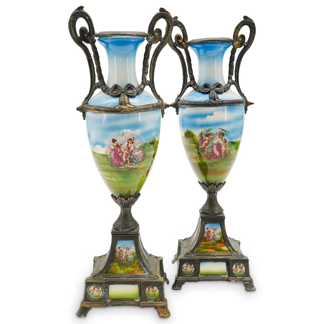 Pair Of Antique French Porcelain And Mixed Metal Amphora Vases: DESCRIPTION: Two matching amphora vases, each composed of porcelain bearing a pair of garland festooned metal handles and mounted to a mixed metal base sporting a porcelain panel. Porcelain components
