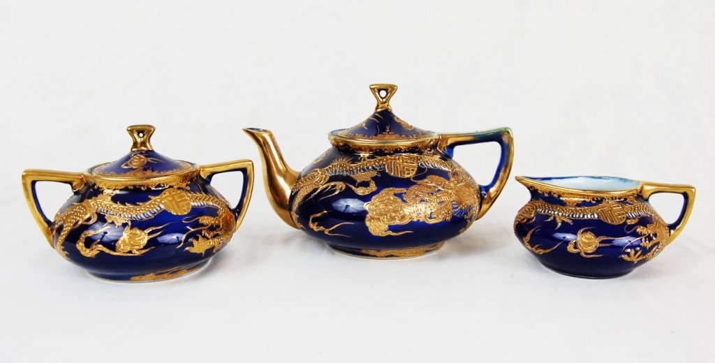 Three piece Japanese Nippon tea set. Hallmarked: DESCRIPTION: Set of three Japanese tea set pieces. The lidded tea pot, lidded sugar bowl, and creamer. White porcelain painted in cobalt blue with a heavy gold hand painted dragon design around each p