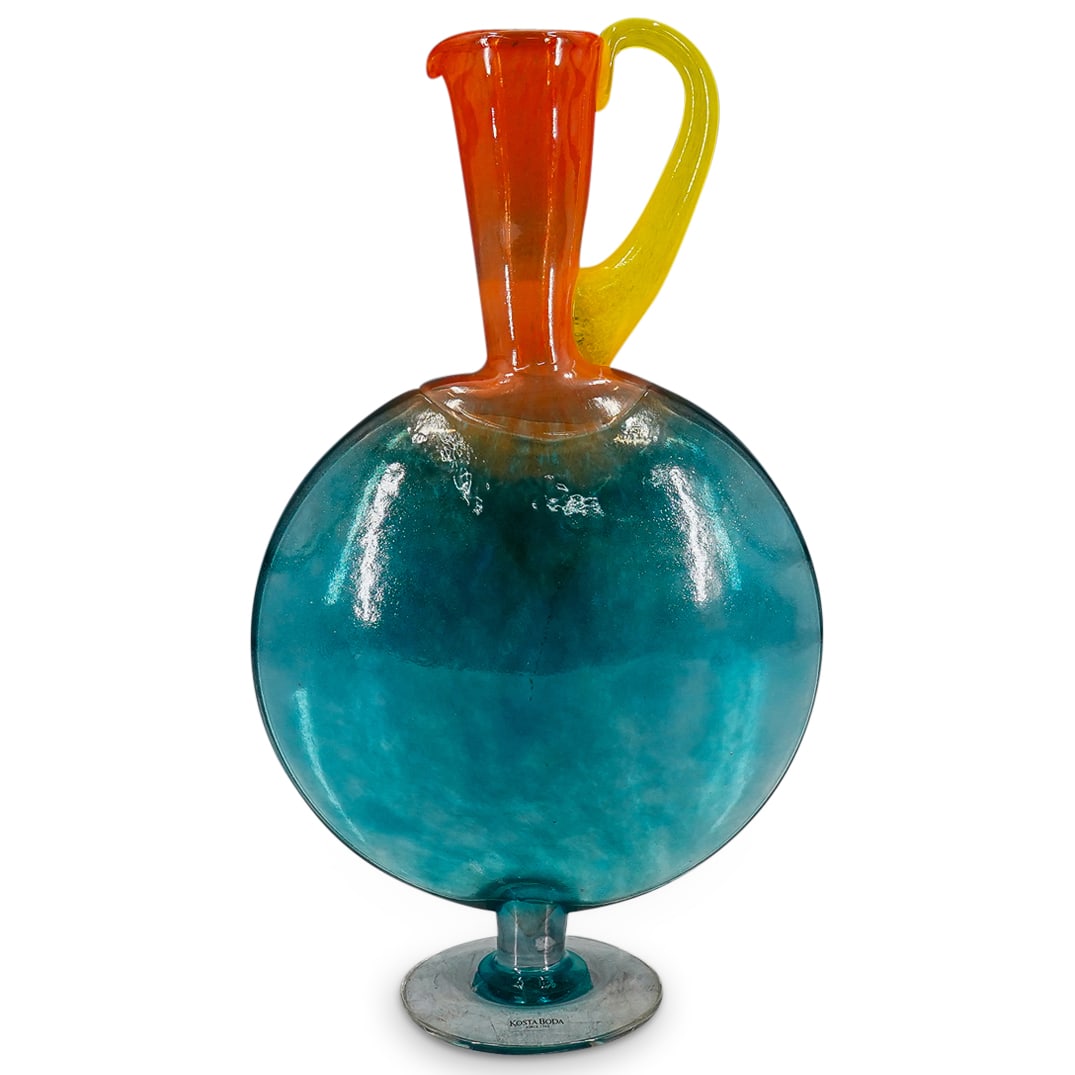 Kosta Boda Glass Flask Form Pitcher: DESCRIPTION: Kosta Boda art glass pitcher featuring a blue, flask form body on a flared foot and sporting an orange neck and lip finished with a yellow handle. CIRCA: 20th Century ORIGIN: Sweden DIMEN