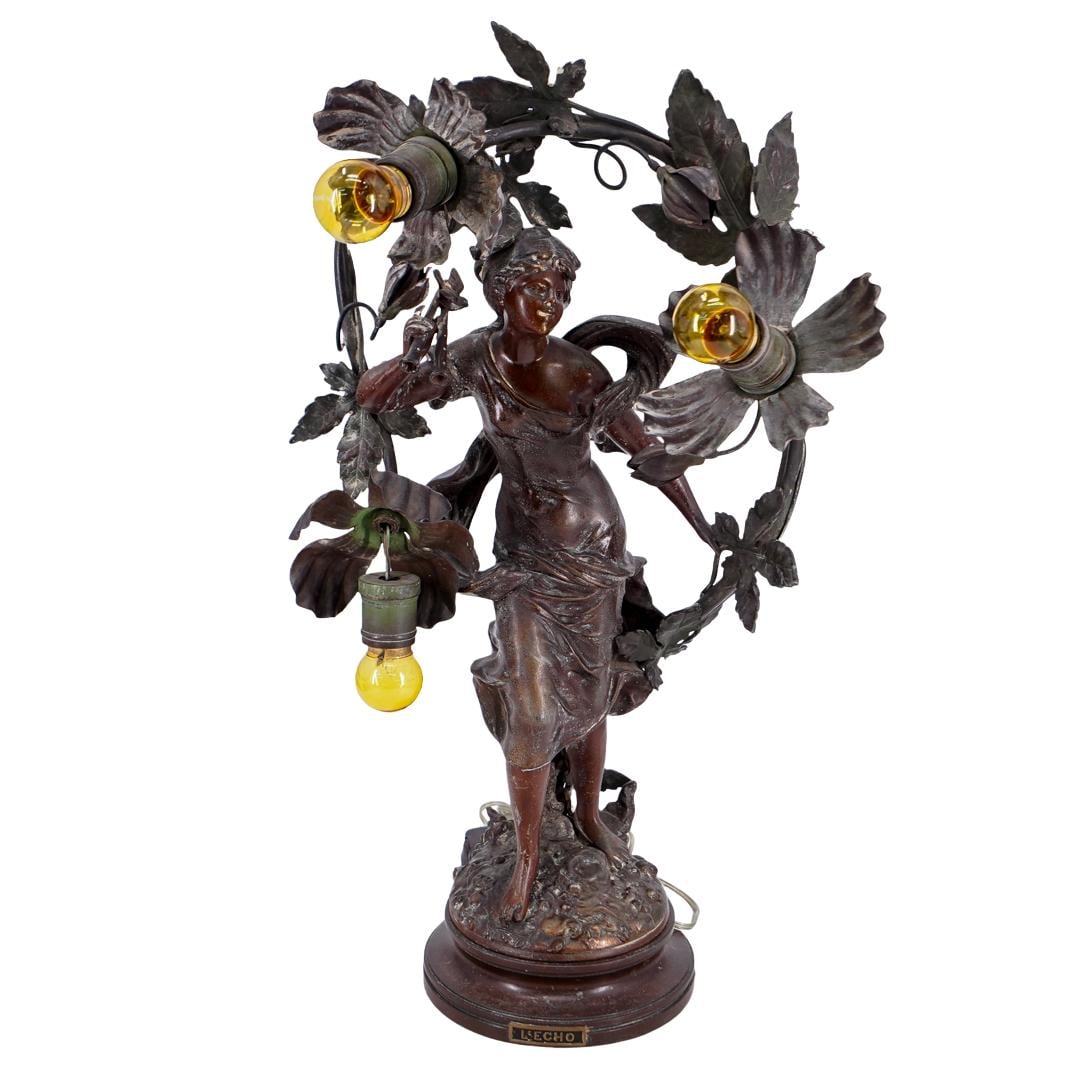 Art Nouveau Wreathed "L'Echo" Bronze Table Lamp: DESCRIPTION: Art Nouveau table lamp crafted as Echo, the nymph, bearing an aulos and standing wreathed with flowers and ivy on a textured base. Plaque branded "L'Echo" present above foot of base. Test