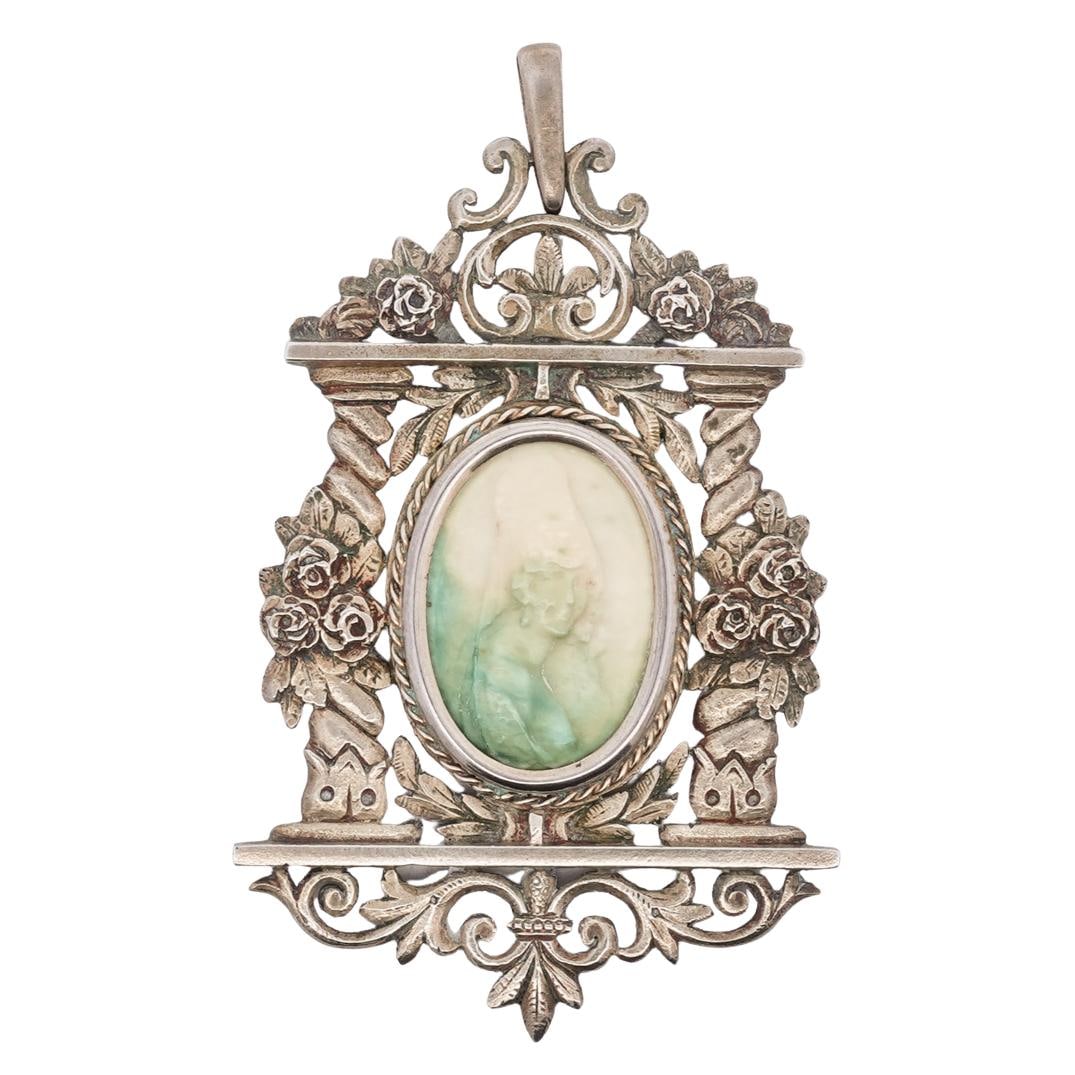 800 Silver Pate de Verre Cameo Pendant: DESCRIPTION: 800 silver cameo pendant, featuring a central oval pate de verre cameo in green and ivory tones. The cameo is surrounded by intricate floral and scrollwork detailing. The top has a decora