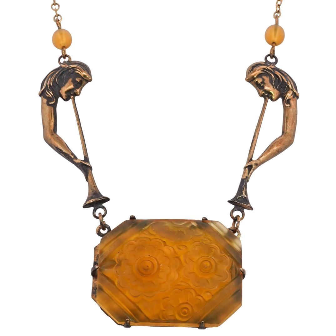 Art Nouveau Amber Glass Necklace: DESCRIPTION: Art nouveau necklace, featuring a fine gold tone chain with two figural figures holding a trumpet on either side that connects to a carved amber camphor glass pendant. CIRCA: 1930's ORIGI