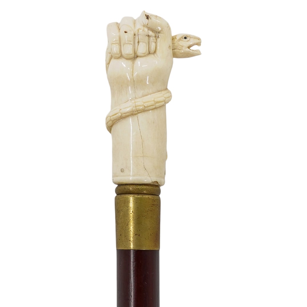 Vintage Wood And Carved Bone Serpent Cane: DESCRIPTION: A vintage cane comprised of a wooden foundation, fitted with a carved bone handle depicting a hand gripping a serpent. CIRCA: Mid-Late 20th Cent. ORIGIN: USA DIMENSIONS: H: 36.5" W: 1.5" 