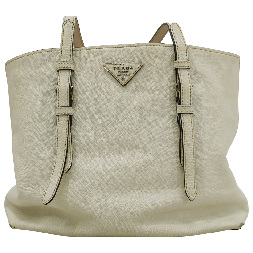 Prada White Saffiano Double Buckle Tote: DESCRIPTION: This Prada tote bag is in white leather with a black trim. Magnetic button closure, and a leather double handle/strap with a silver tone buckle detail on each side, Prada logo in white an