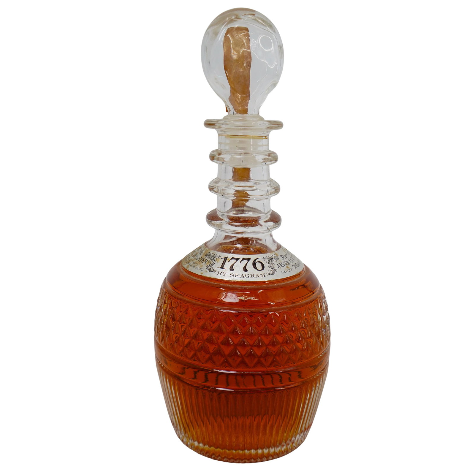 Tiffany Decanter and 1776 Seagram's Premium American Whiskey: DESCRIPTION: This decanter bottle, filled with whiskey, features a textured glass design with a rounded base and a decorative band around the neck. The stopper is made of glass, and a seal strip with 