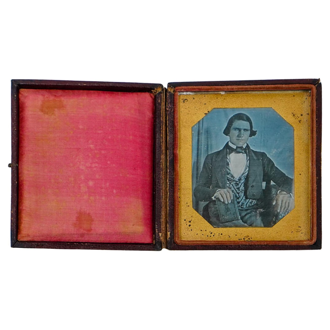 Antique Daguerreotype Portrait Of A Man: DESCRIPTION: Antique daguerreotype portrait, depicting a well dressed man seated in a formal pose. He wear a dark frock coat, waistcoat with a patterned or embroidered design, white shirt, and a bowti